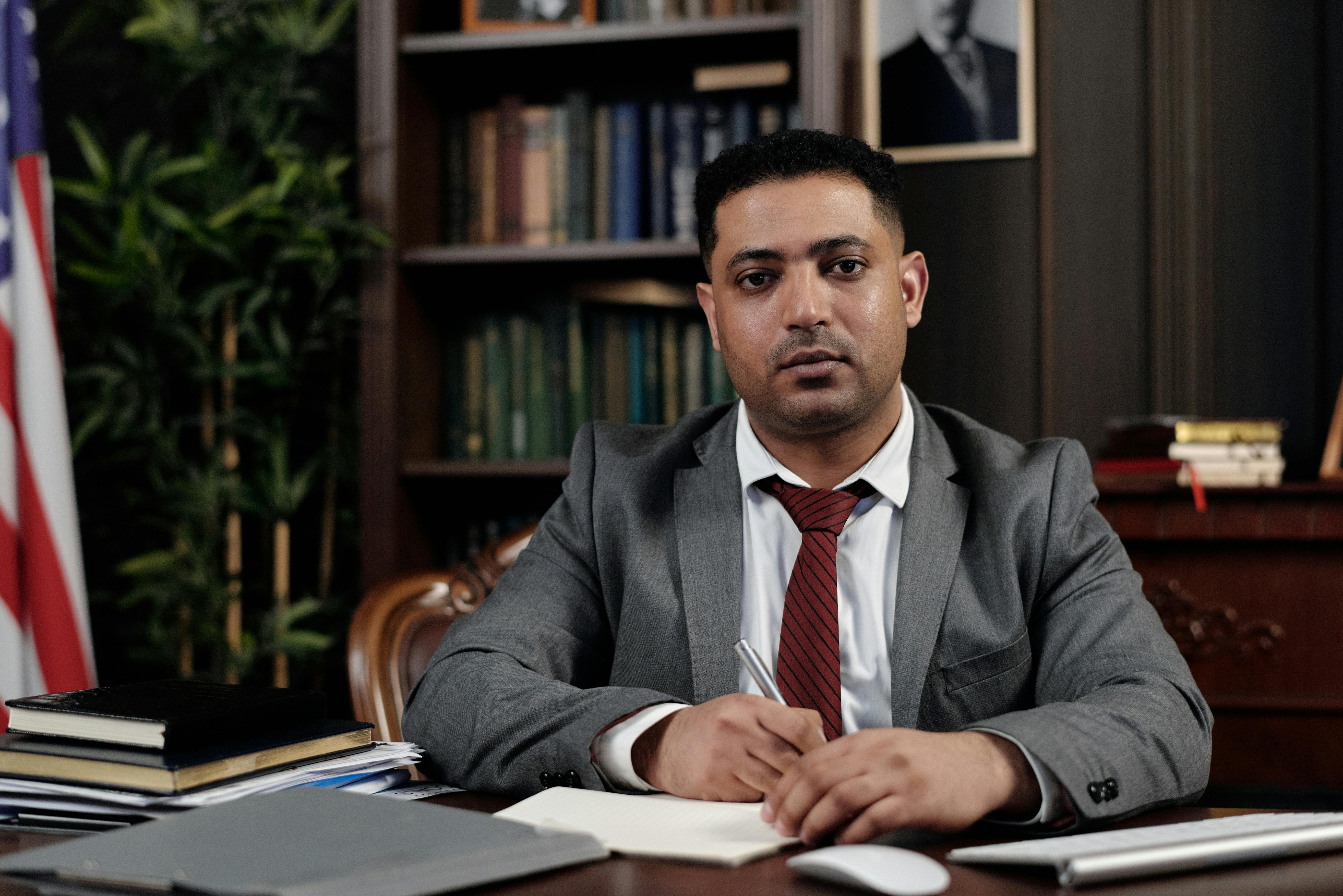 A serious businessman in a suit writing at his desk in a formal office setting.